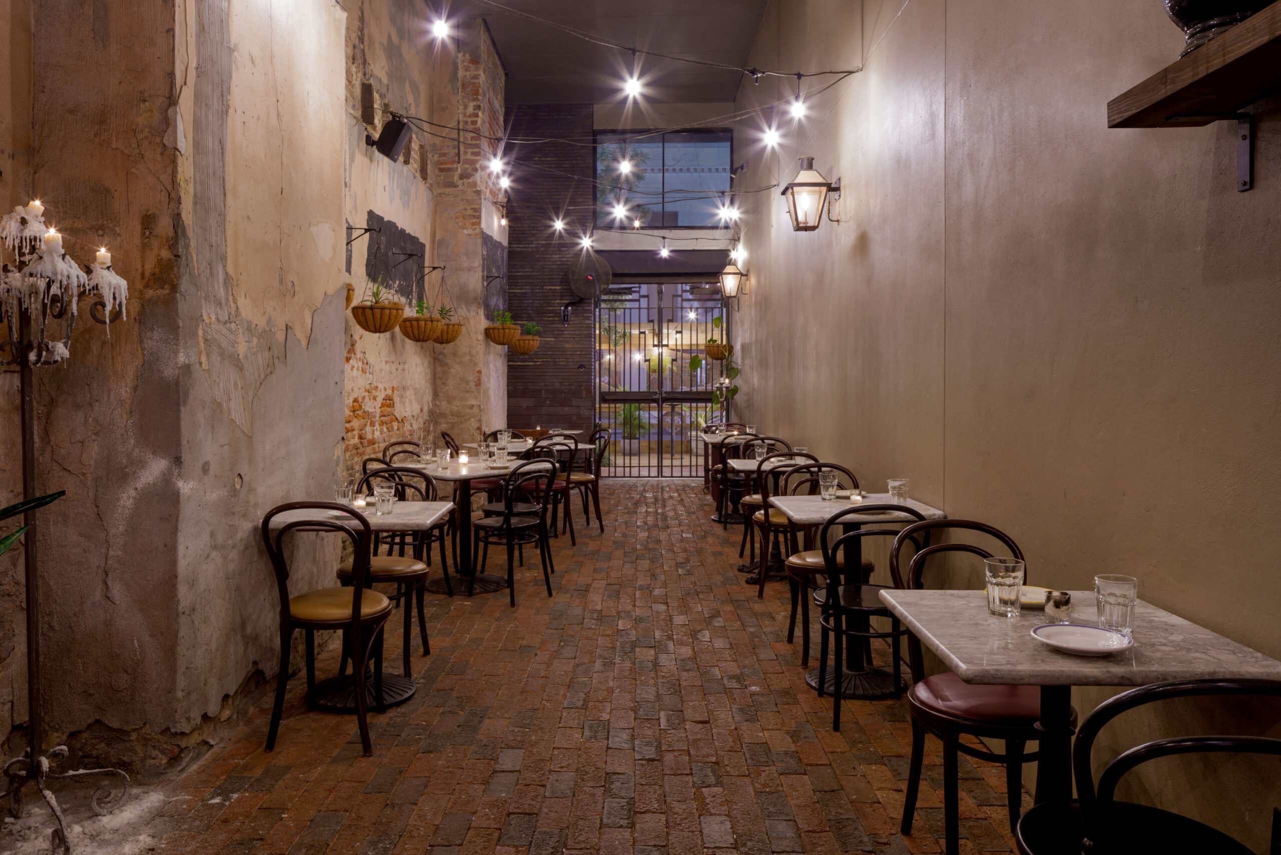 Narrow brick-paved courtyard dining area featuring bistro tables and warm string lighting at night