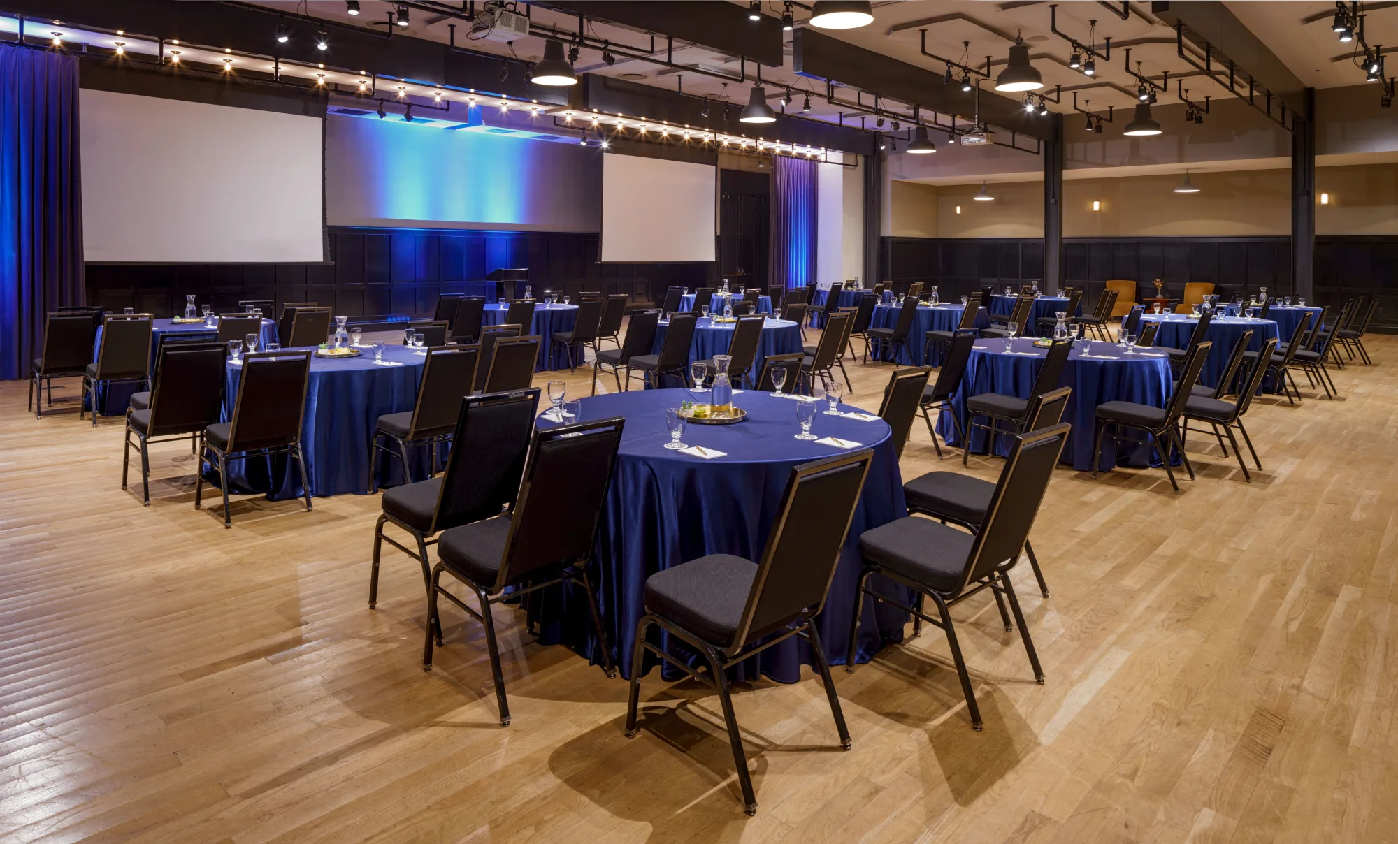 Professional shot of a large ballroom with round blue tables, black chairs, and twin projection screens