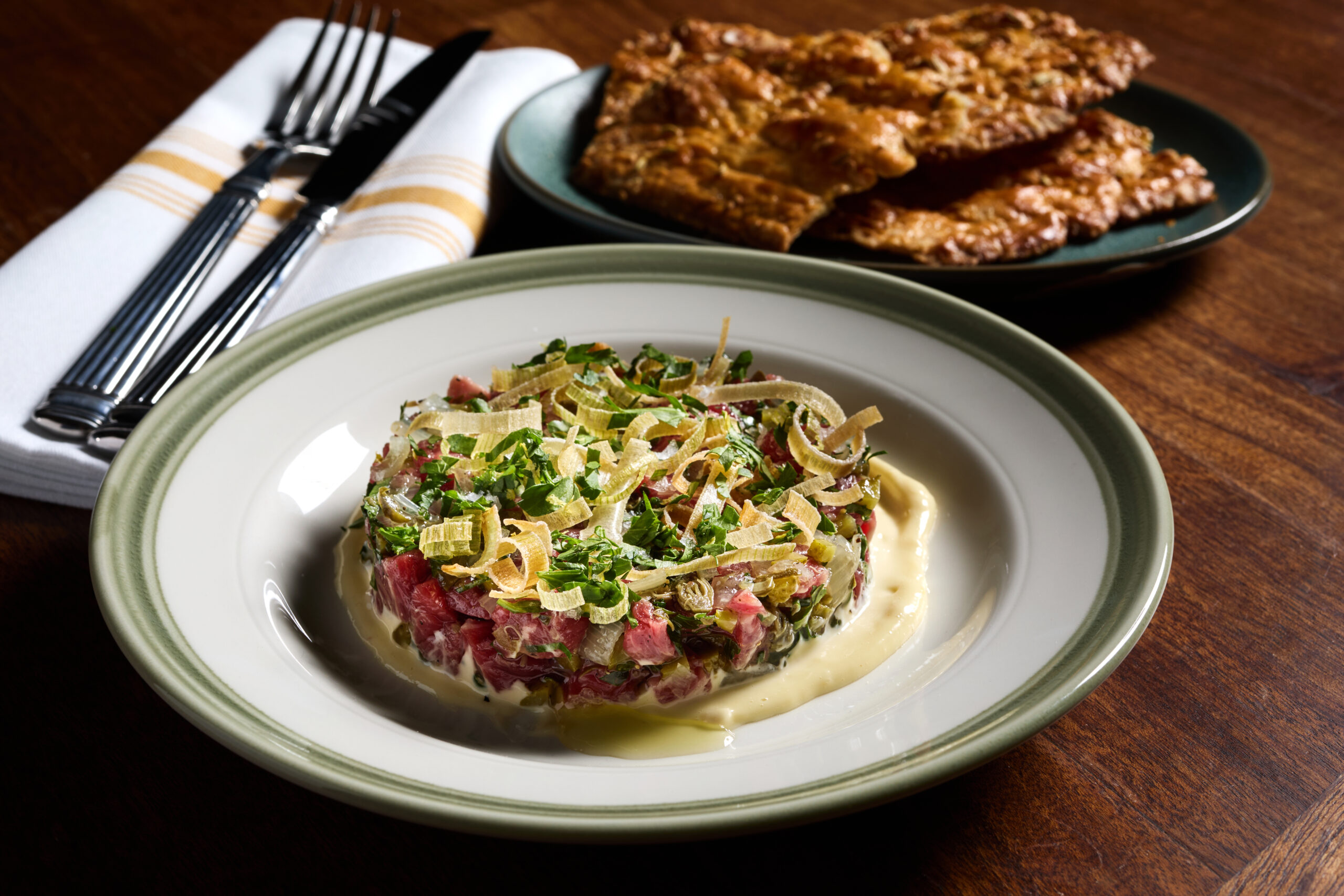 Professional shot of beef tartare garnished with crispy leeks, served with crackers and cutlery