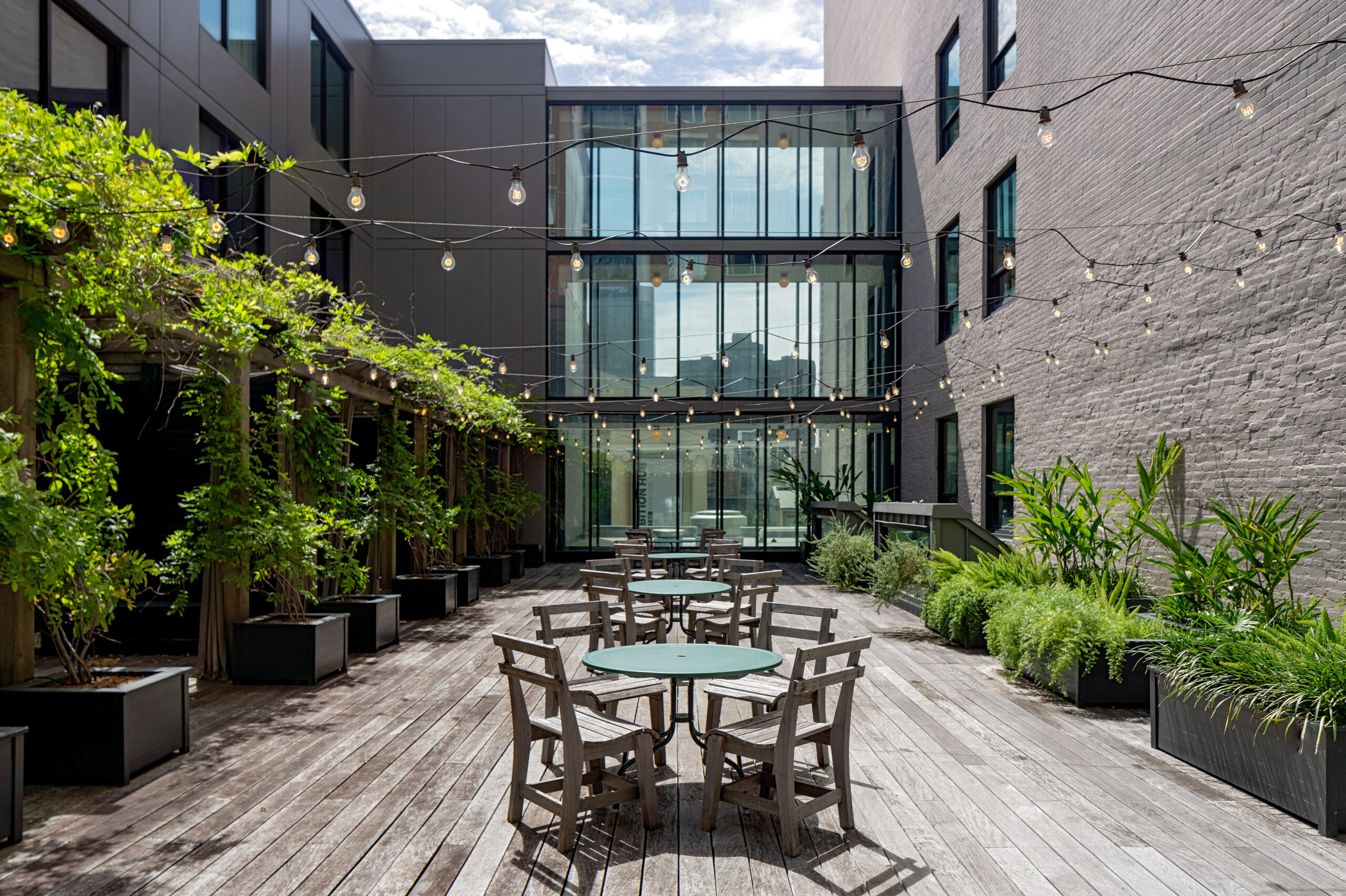Brightly lit wooden deck courtyard with cafe seating, lush plants, and ambient string lights
