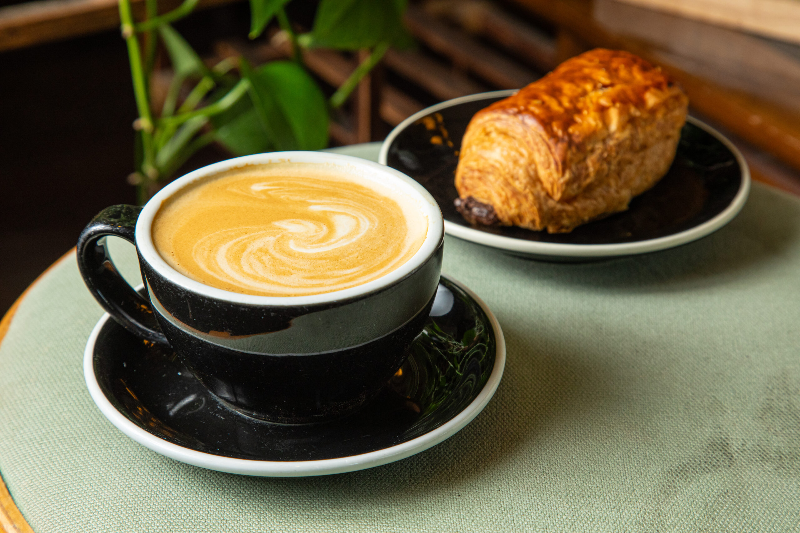 Professional shot of a creamy latte with latte art and a pain au chocolat on a green upholstered surface