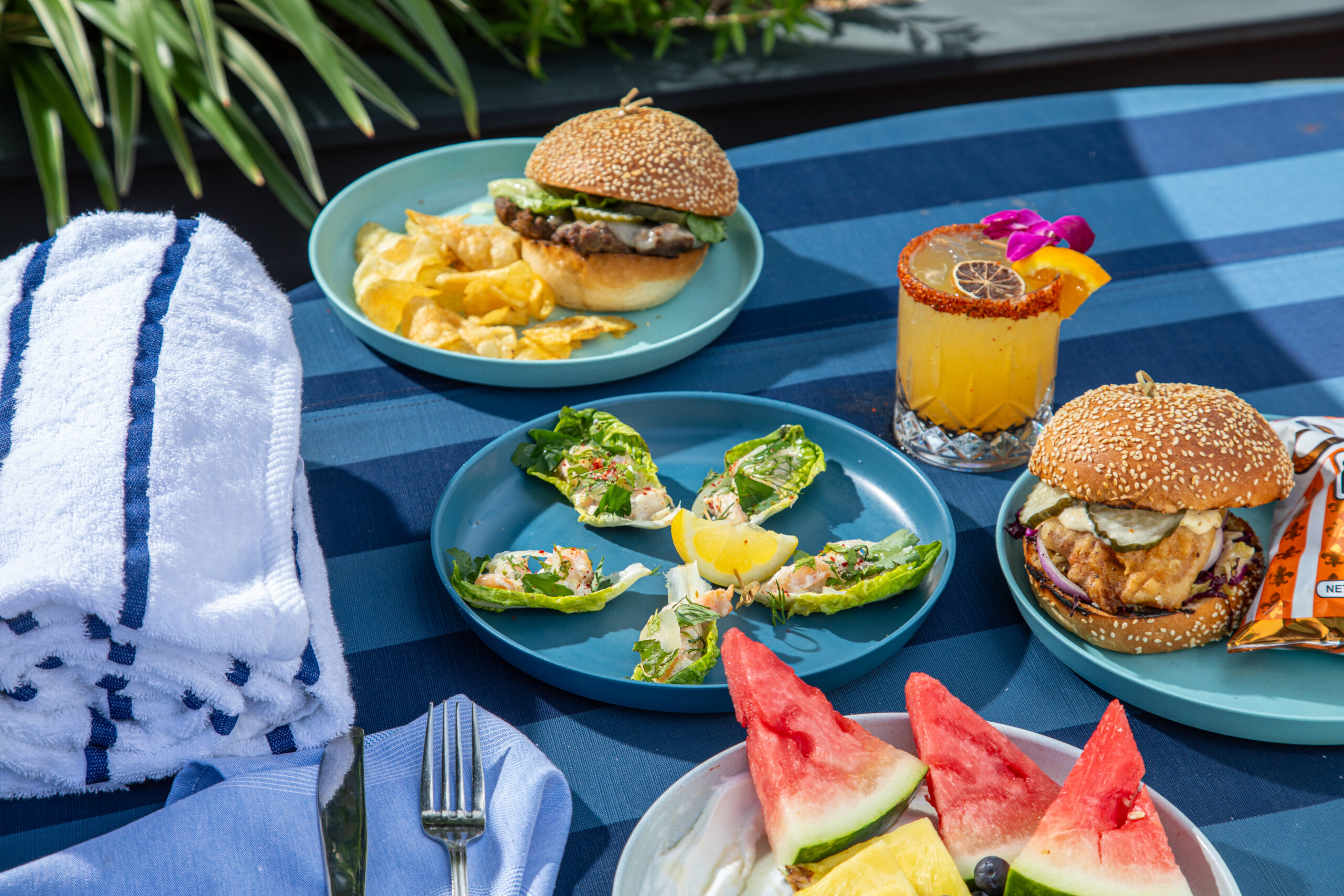 Brightly lit food spread with burgers, tropical cocktail, and fruit salad on a blue striped table