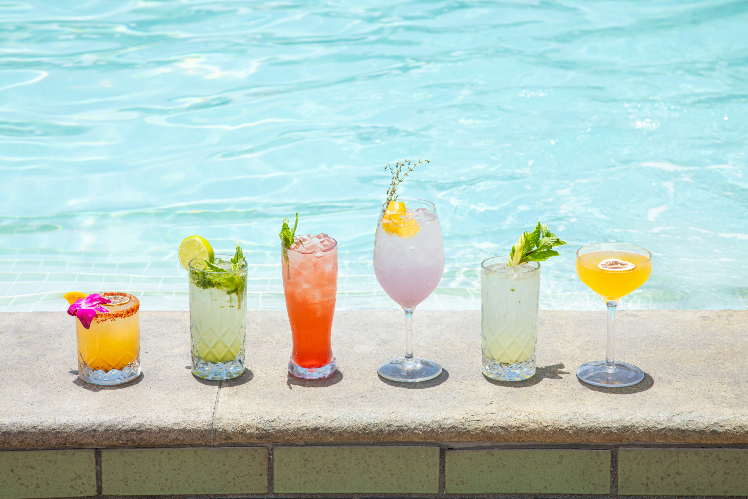 Professional shot showcasing six colorful cocktails resting on the edge of a bright hotel swimming pool